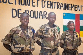 Three young soldiers in camouflage uniforms stand in front of a wall with text and a multicolored flag.