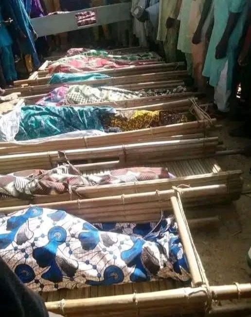 Several covered bamboo stretchers lay in a row, surrounded by onlookers in colorful clothing.