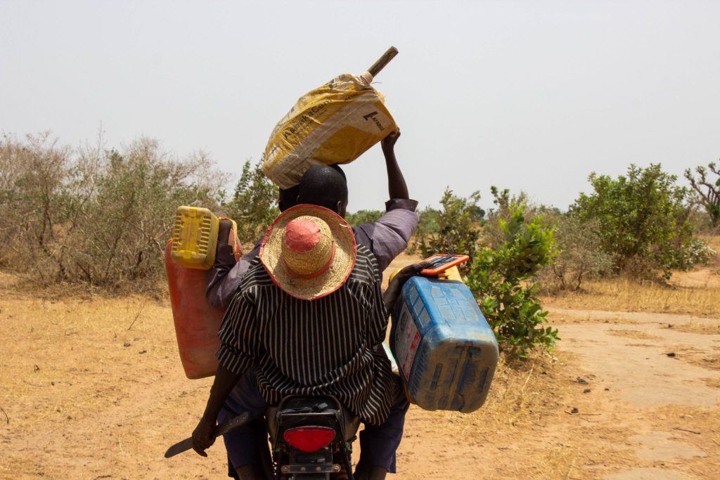 Two people on a motorbike carrying jugs and a sack across a dry rural landscape, one wears a straw hat.