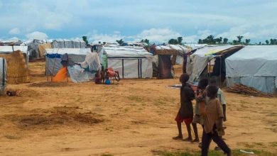 Children walk near makeshift tents in a dry, open area under a cloudy sky.