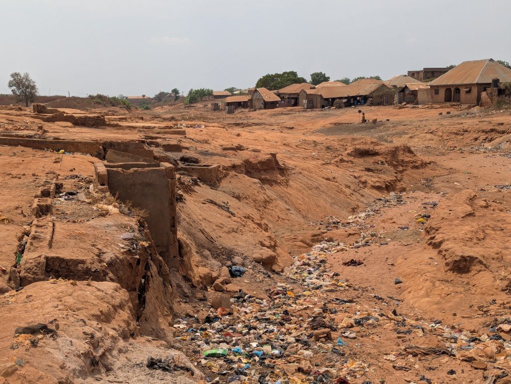 Erosion on arid land with scattered trash and small, distant houses under a hazy sky.