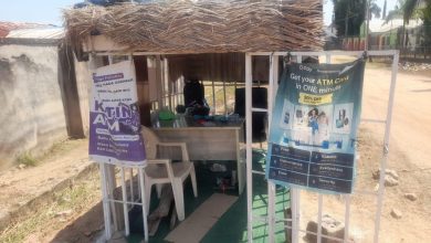 Small outdoor booth with straw roof, advertising ATM and banking services. Two banners hang at the entrance, with chairs and a table inside.
