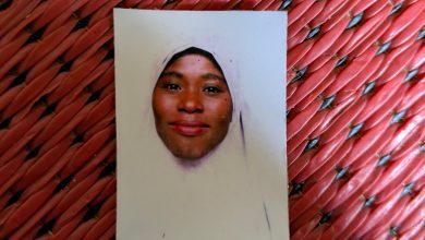 A portrait photo of a person in a light-colored headscarf on a red woven surface.