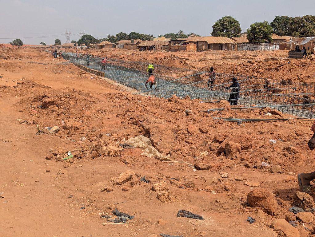 Workers construct a canal with metal reinforcements in a rural area surrounded by red earth and small buildings.