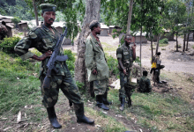 Soldiers in camouflage stand with rifles under trees in a rural setting with wooden buildings in the background.