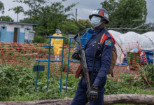 A masked peacekeeper stands with a rifle in a fenced area, while a person in protective gear walks in the background.