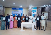Group of people at a book launch event, each holding a copy of the book. Banners and a table with a white cloth are in the background.